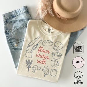 May include: Ivory-colored t-shirt with a baking-themed graphic. The design includes illustrations of a rolling pin, whisk, bread, chef's hat, and the words "flour water salt." The shirt is paired with light blue jeans and a beige hat.