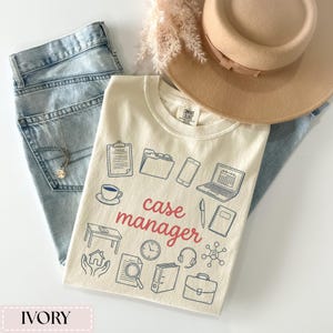 May include: Ivory-colored t-shirt with the words "case manager" in red script, surrounded by blue line drawings of work-related items. The shirt is paired with light-wash jeans, a tan hat, and a necklace.