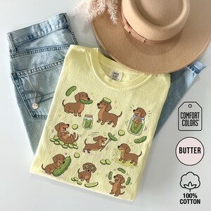 May include: A light yellow Comfort Colors t-shirt featuring cartoon dachshunds with pickles. The shirt has a crew neck and short sleeves. The shirt is paired with blue jeans and a tan hat. The shirt is made of 100% cotton.