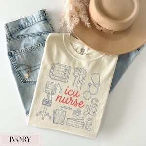 May include: Ivory-colored t-shirt with blue medical equipment illustrations and the words "icu nurse" in red. The shirt is paired with light blue jeans, a tan hat, and a necklace. The shirt is made by Comfort Colors.