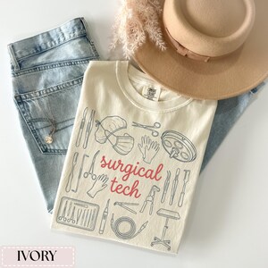 May include: Ivory-colored t-shirt with a "surgical tech" design in red and blue. The shirt features illustrations of surgical tools, a surgical cap, mask, gloves, and a surgical lamp. A light brown hat and light blue jeans are also in the image.