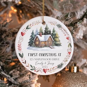 May include: A round white ceramic ornament featuring a central illustration of a snow-covered cabin surrounded by pine trees. The text reads FIRST CHRISTMAS AT 123 MAPLEWOOD DRIVE Emily & Jessy 2025. A decorative wreath of red hearts and green leaves borders the design.