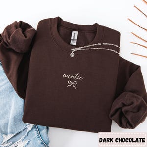 May include: A dark chocolate-colored sweatshirt with the word "auntie" embroidered in white script, along with a bow graphic. A silver necklace with a sun pendant rests on the sweatshirt. The text "DARK CHOCOLATE" is at the bottom.