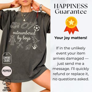 May include: Dark grey Comfort Colors t-shirt with the phrase "outnumbered by boys" and various white graphics including a truck, football, baseball, soccer ball, robot, dinosaur, and tools. The shirt is made of 100% cotton.