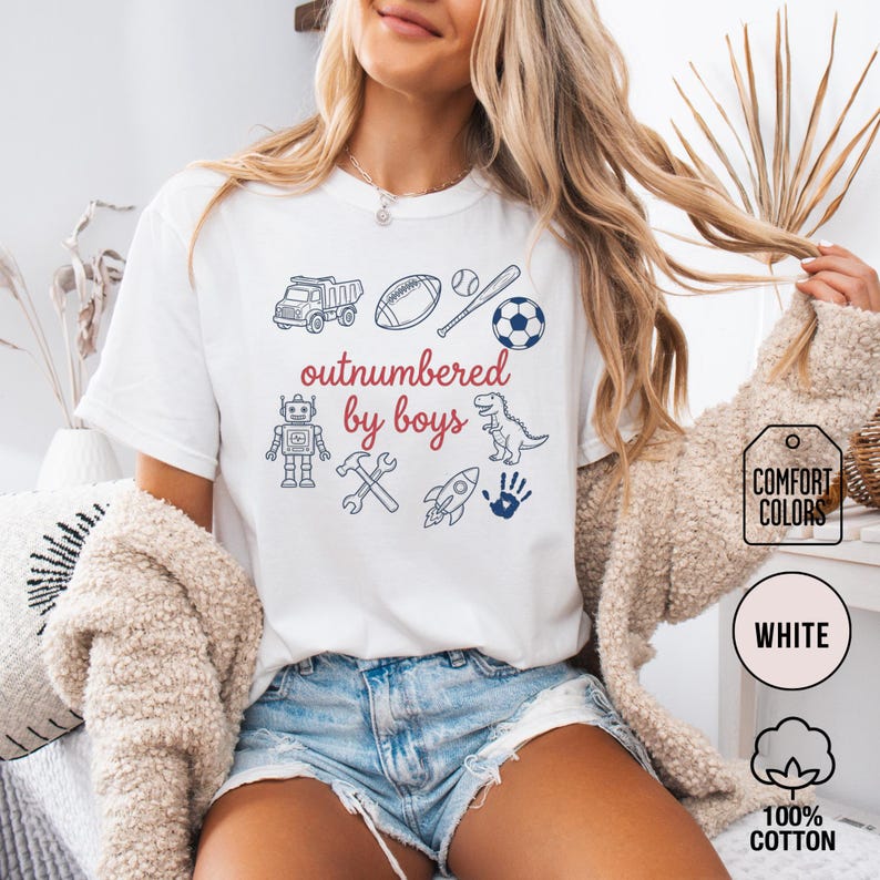 May include: White t-shirt with the phrase "outnumbered by boys" in red and blue, accompanied by illustrations of a truck, football, baseball, soccer ball, robot, hammer, wrench, rocket, dinosaur, and handprint. The shirt is labeled "Comfort Colors" and made of 100% cotton.