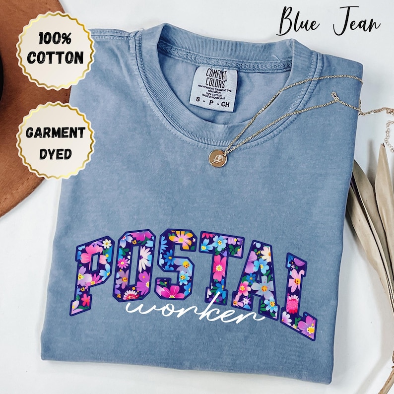 May include: Blue jean coloured t-shirt with the words "POSTAL worker" in a floral design. The shirt is made of 100% cotton and is garment dyed. A gold necklace with a pendant is on the shirt.