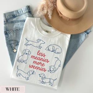 May include: White t-shirt with a crew neck featuring blue line drawings of dachshunds and the text "less meanies more weenies" in red. The shirt is paired with light blue jeans and a tan hat.