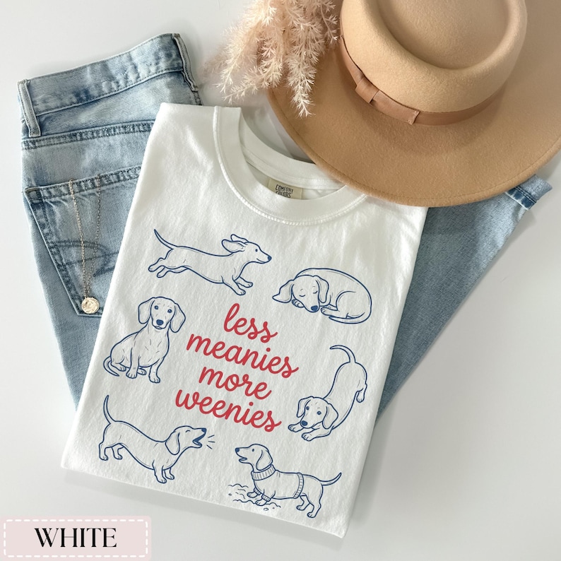 May include: White t-shirt with a crew neck featuring blue line drawings of dachshunds and the text "less meanies more weenies" in red. The shirt is paired with light blue jeans and a tan hat.