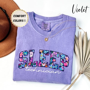 May include: A violet Comfort Colors® t-shirt with the word "SLEEP" in floral letters and the word "technician" in white script. A gold necklace and a brown hat are also in the image.