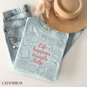 May include: A light blue t-shirt with the text "Life happens visuals help" and various graphic icons. The shirt is paired with light blue jeans, a tan hat, and a gold necklace. The word "CHAMBRAY" is at the bottom.
