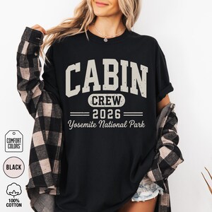 May include: A black t-shirt featuring the text CABIN CREW 2026 Yosemite National Park in a distressed beige font. The shirt is layered under a black and beige plaid flannel shirt. The design includes a Comfort Colors tag and 100% cotton label.