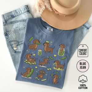 May include: Blue jean Comfort Colors t-shirt with a playful illustration of dachshunds and pickles. The shirt features multiple brown dachshunds interacting with pickles and jars of pickles. The shirt is made of 100% cotton.