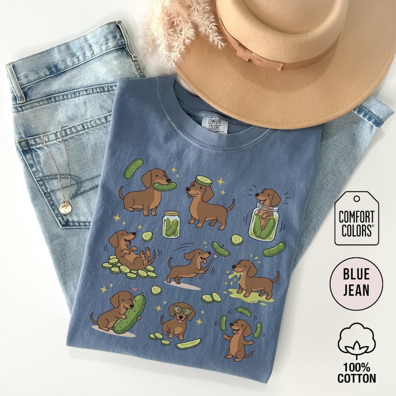 May include: Blue jean Comfort Colors t-shirt with a playful illustration of dachshunds and pickles. The shirt features multiple brown dachshunds interacting with pickles and jars of pickles. The shirt is made of 100% cotton.