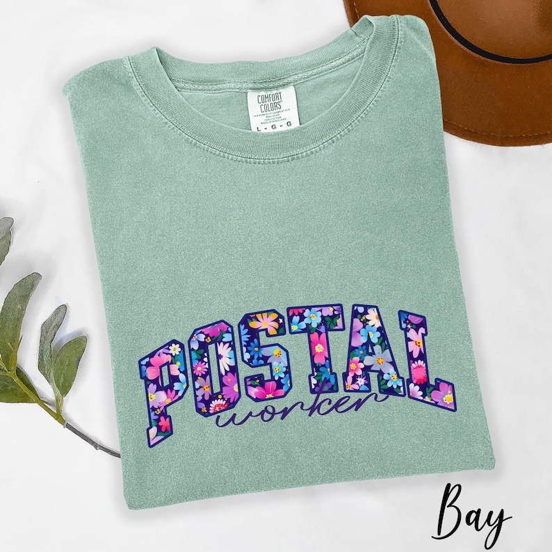 May include: A sage green t-shirt featuring the words "POSTAL worker" in a floral design. The shirt has a crew neck and is made of a soft material. The brand "Comfort Colors" is visible on the tag.