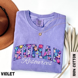 May include: A violet Comfort Colors t-shirt with "HUMAN Resources" in a floral design. The shirt has a crew neck and is made of 100% cotton. A gold necklace with a pendant rests on the shirt. The word "VIOLET" is at the bottom left.
