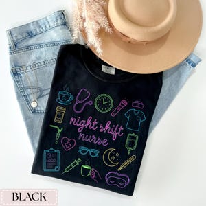 May include: Black t-shirt with neon illustrations of medical symbols and the words "night shift nurse". The shirt is paired with light blue jeans and a tan hat. A gold necklace is visible in the jeans pocket.