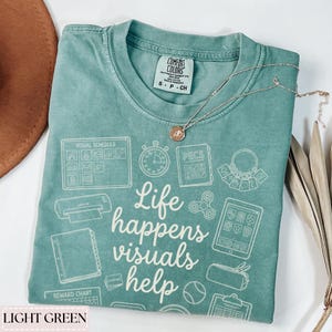 May include: Light green t-shirt with the text "Life happens visuals help" and various visual aid graphics. The shirt has a crew neck and short sleeves. A gold necklace with a pendant is draped across the shirt.