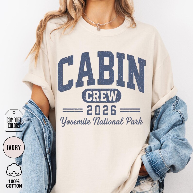 May include: An ivory short-sleeve t-shirt featuring dark blue distressed lettering that reads CABIN CREW 2026 Yosemite National Park. The shirt has a crew neckline and is layered under a blue denim jacket, showcasing a casual and relaxed fit.