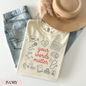 May include: Ivory-colored t-shirt with the phrase "your words matter" in red script, surrounded by line drawings of a microphone, brain, and other communication-related symbols. The shirt is paired with light-wash jeans, a necklace, and a tan hat.