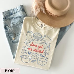 May include: Ivory-colored t-shirt with a blue and red graphic of baking tools and the phrase "don't get me started." The shirt is paired with light blue jeans, a tan hat, and a gold necklace. The shirt is made of a soft material.