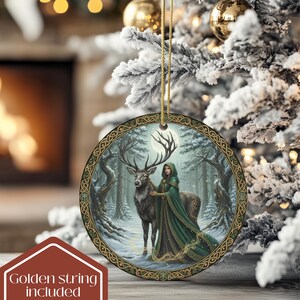 May include: Round Christmas ornament with a winter forest scene. The ornament features a woman in a green cloak with a deer, set against a snowy forest backdrop. The ornament has a gold border and a golden string for hanging. The text "Golden string included" is in the lower left corner.