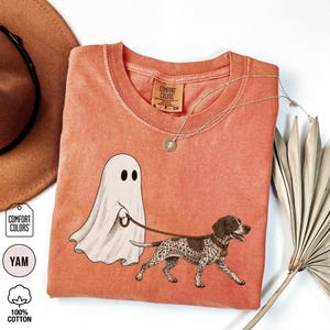 May include: A burnt orange short-sleeve t-shirt featuring a graphic of a white ghost walking a brown and white spotted dog on a leash. The shirt has a crew neckline and a Comfort Colors brand label printed on the inside of the collar area.
