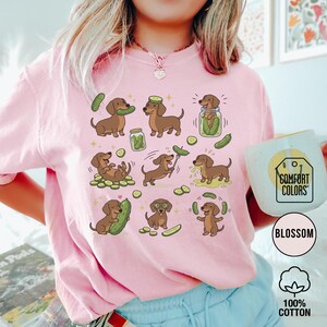 May include: Pink Comfort Colors sweatshirt with a repeating pattern of brown dachshunds and pickles. The dachshunds are in various poses, interacting with pickles. The sweatshirt has the text "Blossom" and "100% Cotton" on the bottom right.