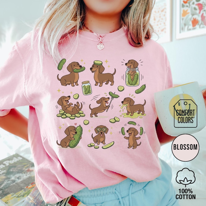 May include: Pink Comfort Colors sweatshirt with a repeating pattern of brown dachshunds and pickles. The dachshunds are in various poses, interacting with pickles. The sweatshirt has the text "Blossom" and "100% Cotton" on the bottom right.