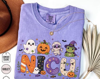 May include: A violet t-shirt featuring a graphic of a ghost, a pumpkin, a mummy, and a frog wearing a witch hat above the text NICU. The letters are decorated with Halloween patterns including candy corn, spiderwebs, and stars. The shirt has a Comfort Colors label.