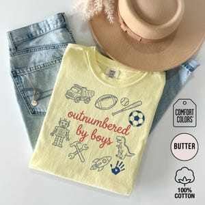 May include: A light yellow Comfort Colors t-shirt with the phrase "outnumbered by boys" in red script. The shirt features blue illustrations of a truck, football, baseball, soccer ball, robot, dinosaur, and other boy-themed icons. The shirt is paired with blue jeans and a tan hat.