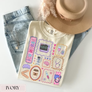 May include: Ivory-colored t-shirt with colorful ICU nurse-themed graphics, including a heart monitor, IV drip, and stethoscope. The shirt is laid on top of light blue jeans, a necklace, and a beige hat.