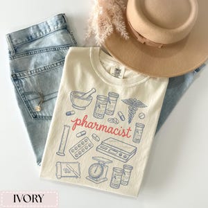 May include: Ivory-colored t-shirt with a pharmacist design. The shirt features illustrations of pharmacy tools and the word "pharmacist" in red script. The shirt is paired with light-wash jeans, a tan hat, and a necklace.