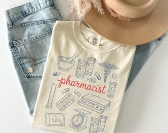 Könnte beinhalten: Elfenbeinfarbenes T-Shirt mit einem Apotheker-Design. Das Shirt zeigt Illustrationen von Apothekenwerkzeugen und das Wort "Apotheker" in roter Schrift. Das Shirt wird mit hellblauen Jeans, einem beigen Hut und einer Halskette kombiniert.