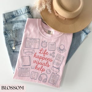 May include: A light pink Comfort Colors t-shirt with the text "Life happens visuals help" and various visual aid icons. The shirt is paired with light wash jeans and a tan hat. A gold necklace is visible in the jeans pocket.