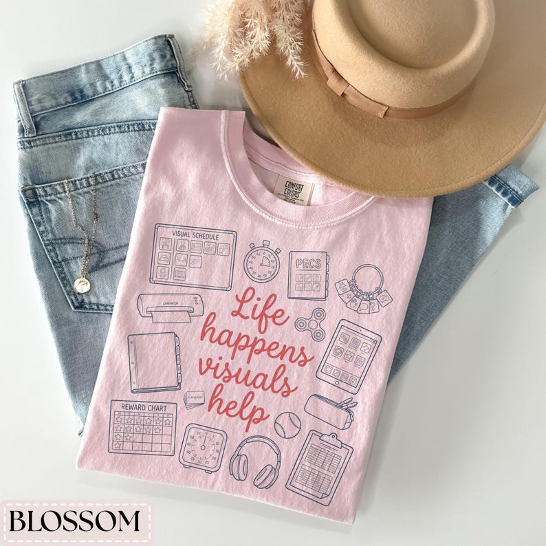 May include: A light pink Comfort Colors t-shirt with the text "Life happens visuals help" and various visual aid icons. The shirt is paired with light wash jeans and a tan hat. A gold necklace is visible in the jeans pocket.