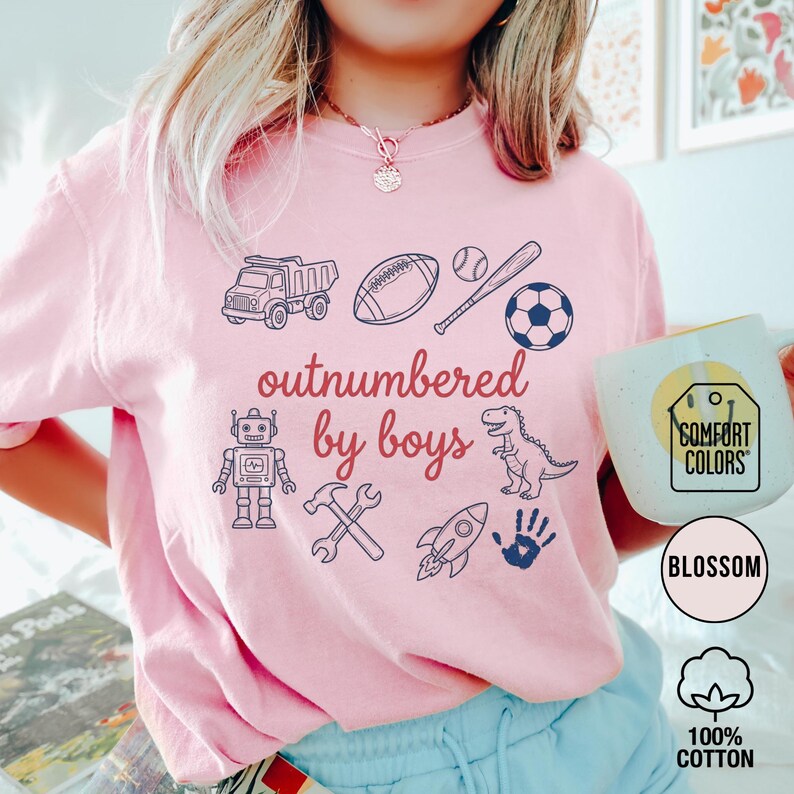 May include: A light pink t-shirt with the text "outnumbered by boys" and various illustrations, including a truck, football, baseball, soccer ball, robot, hammer, wrench, rocket, dinosaur, and handprint. The shirt is made of 100% cotton.