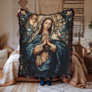 May include: A woven throw blanket featuring a religious image of a woman with a halo, hands clasped in prayer, and wearing a blue robe. The blanket has a fringed edge with red and black accents. The artwork resembles a stained-glass window.