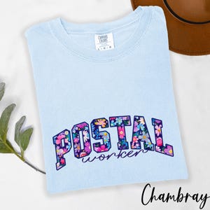 May include: Pale blue t-shirt featuring the text "POSTAL worker" in a floral design. The shirt has a crew neck and the words "Comfort Colors" are printed on the upper left side. The shirt is a light blue colour.