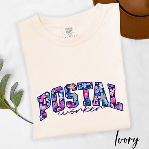 May include: Ivory t-shirt featuring the text "POSTAL worker" in a floral design. The text is in a dark blue font with a floral pattern of pink, purple, and blue flowers. The shirt is made by Comfort Colors.