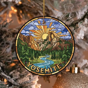 May include: Round Christmas ornament featuring a stained-glass design of Yosemite National Park. The image includes a river, mountains, trees, and the text "YOSEMITE". The ornament is trimmed in gold and hangs from a gold chain.