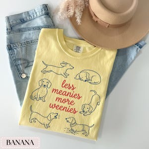 May include: A yellow t-shirt with a dachshund dog graphic and the text "less meanies more weenies". The shirt is paired with blue jeans and a tan hat. The shirt is a banana color.
