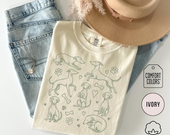 May include: Ivory-colored t-shirt with a light green dog illustration. The shirt features various dog poses, paw prints, hearts, and bones. The shirt is made of 100% cotton and is paired with light blue jeans and a tan hat.