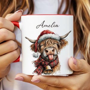 May include: White ceramic mug with a red interior and handle. The mug features a watercolor illustration of a Highland cow wearing a Santa hat and scarf. The name "Amelia" is written above the cow.