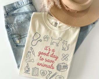 May include: Ivory-colored t-shirt with the phrase "it's a good day to save animals" and various veterinary-themed illustrations. The shirt is paired with light-wash denim jeans and a beige hat.