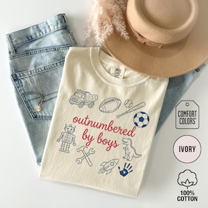 May include: Ivory-colored t-shirt with the text "outnumbered by boys" in red and blue graphics of a truck, football, baseball, soccer ball, robot, hammer, wrench, rocket, dinosaur, and handprint. The shirt is made of 100% cotton.