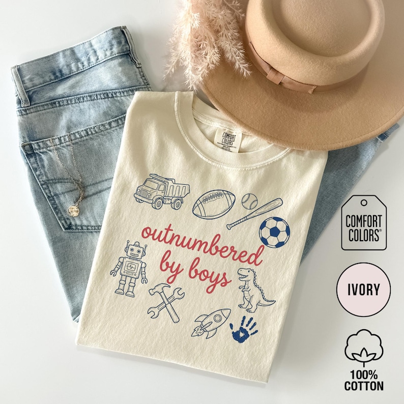 May include: Ivory-colored t-shirt with the text "outnumbered by boys" in red and blue graphics of a truck, football, baseball, soccer ball, robot, hammer, wrench, rocket, dinosaur, and handprint. The shirt is made of 100% cotton.