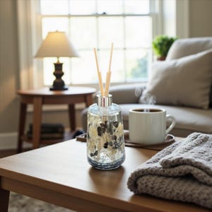 May include: A clear glass essential oil diffuser with silver accents and wooden reeds. The diffuser sits on a wooden table next to a white mug of coffee and a folded gray blanket. A lamp and a couch are in the background.