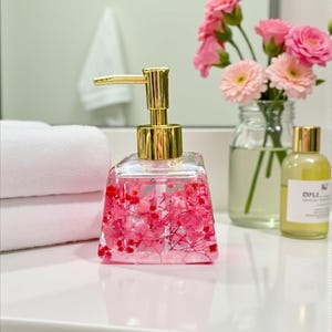 Op de afbeelding: Een heldere, vierkante zeepdispenser met een gouden pomp, gevuld met roze bloemen en rode accenten. De dispenser staat op een wit oppervlak, naast witte handdoeken en een kleine vaas met roze bloemen. Een klein flesje vloeistof is ook zichtbaar.