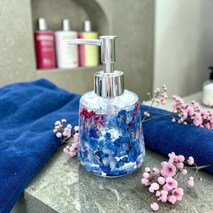 May include: A clear glass soap dispenser with a silver pump mechanism, filled with a liquid containing embedded blue hydrangea petals and small pink flowers. The dispenser has a wide base and sits near a dark blue towel and sprigs of baby's breath.