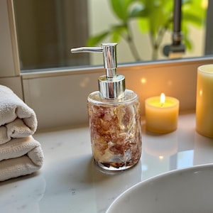 Könnte beinhalten: Ein klarer Seifenspender aus Glas mit silberner Pumpenoberseite. Der Spender ist mit getrockneten Blumen in Braun- und Cremetönen gefüllt. Eine brennende Kerze und gerollte weiße Handtücher befinden sich im Hintergrund.